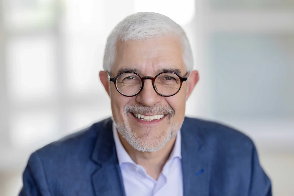 Ein lächelnder älterer Mann mit kurzen grauen Haaren, Brille und einem gepflegten Bart, der einen blauen Blazer und ein helles Hemd trägt und in einem Raum sitzt, dessen Hintergrund leicht unscharf ist.